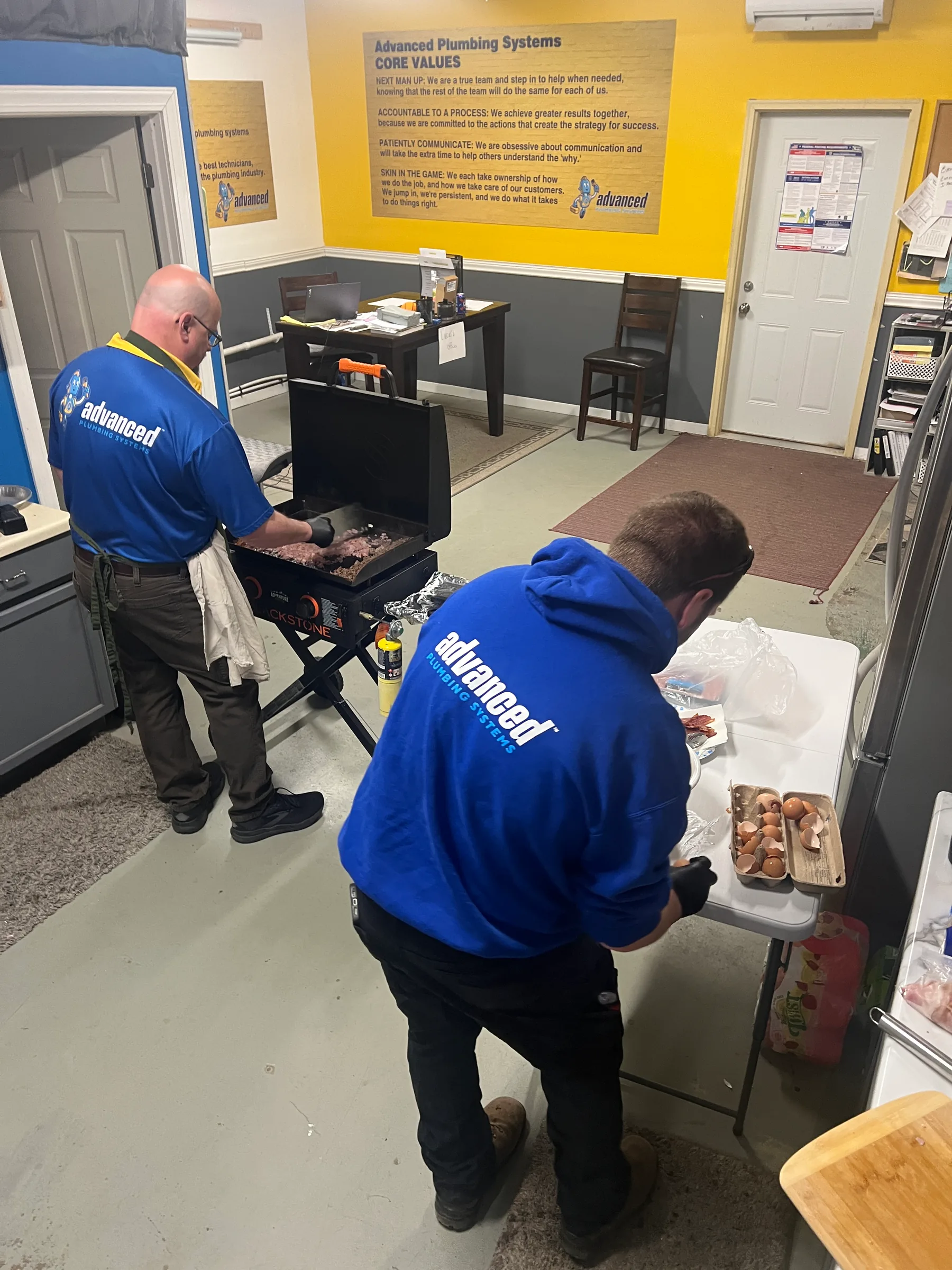 Two Advanced Plumbing Systems employees in blue shirts grilling food at an office event.